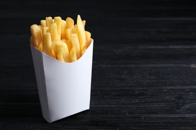 Photo of Delicious french fries in paper box on black wooden table, space for text