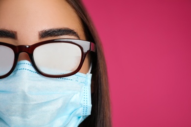 Photo of Young woman with foggy glasses caused by wearing disposable mask on pink background, space for text. Protective measure during coronavirus pandemic