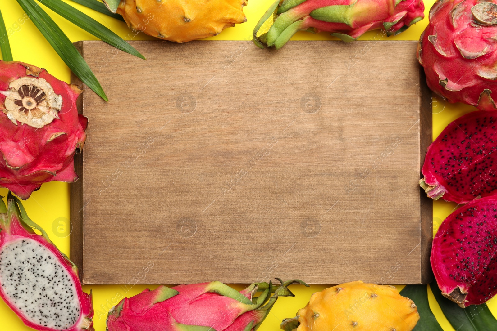 Photo of Frame of different pitahaya fruits, tropical leaves and wooden board on yellow background, flat lay. Space for text