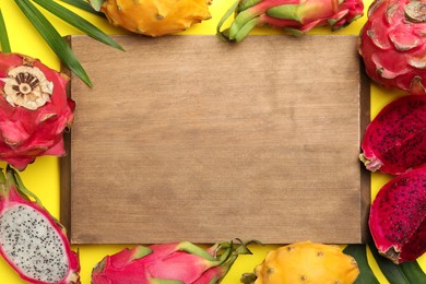 Frame of different pitahaya fruits, tropical leaves and wooden board on yellow background, flat lay. Space for text