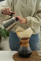 Woman pouring hot water into glass chemex coffeemaker with paper filter and coffee at table, closeup