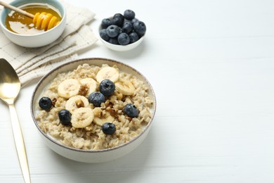 Photo of Tasty oatmeal with banana, blueberries, walnuts and honey served in bowl on white wooden table, space for text