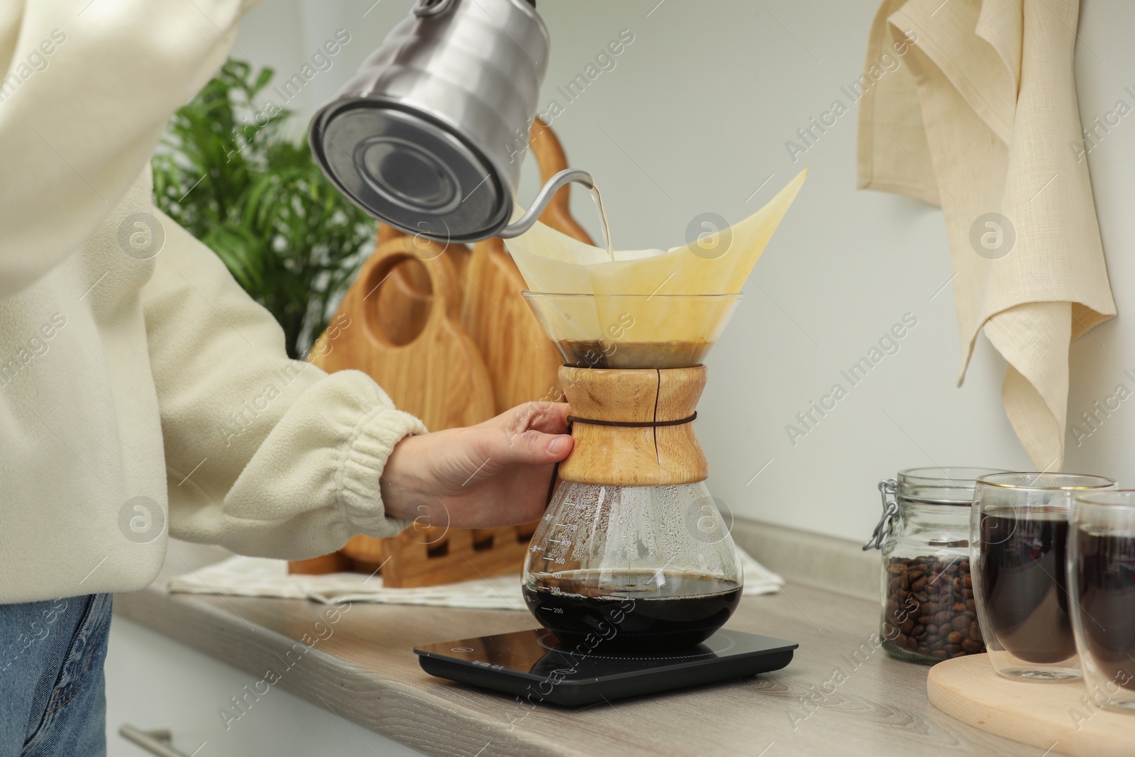 Photo of Woman pouring hot water into glass chemex coffeemaker with paper filter and coffee at countertop in kitchen, closeup