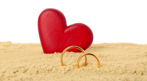 Photo of Honeymoon concept. Two golden rings, red wooden heart and sand isolated on white