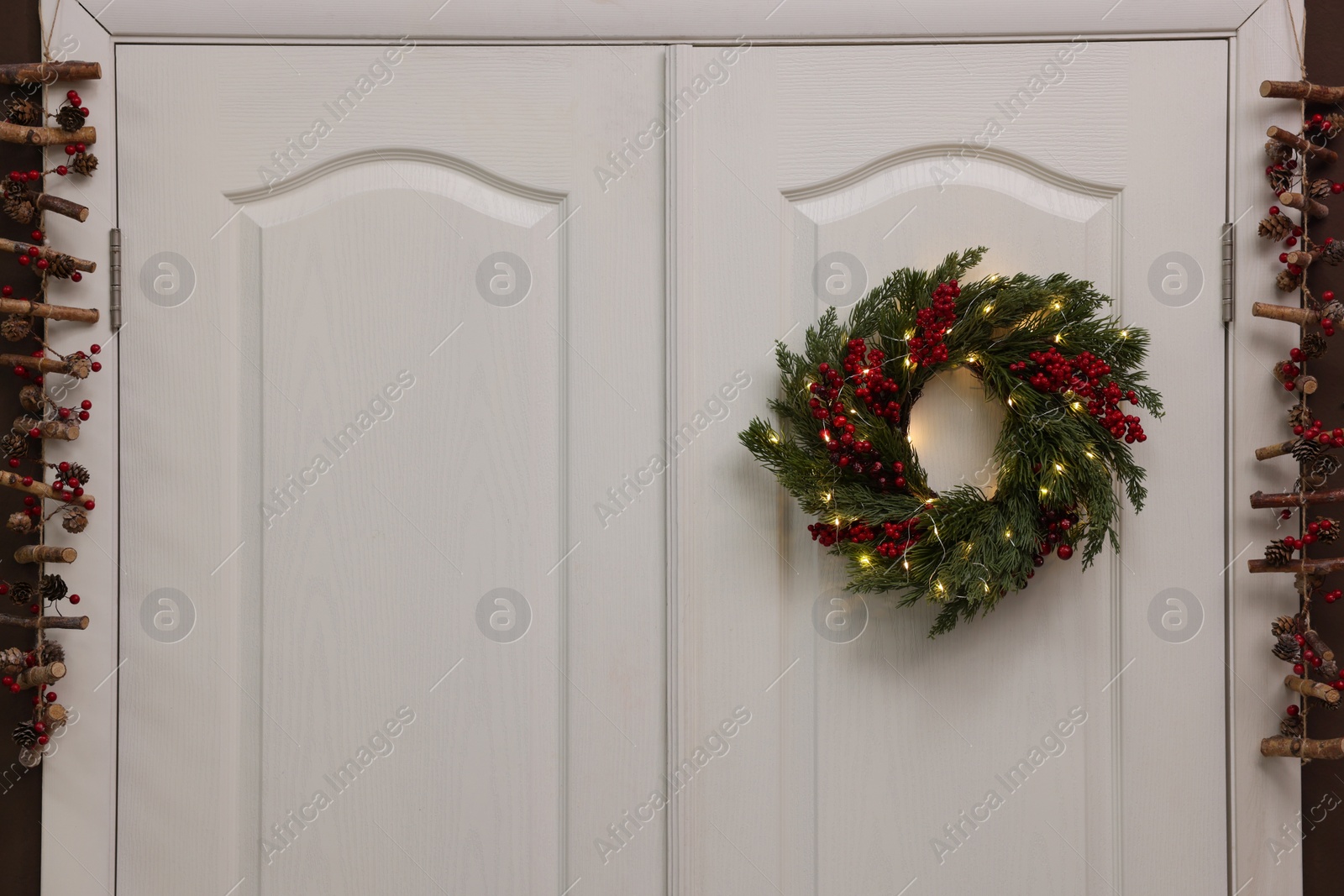 Photo of Beautiful Christmas wreath with red berries and fairy lights hanging on white door