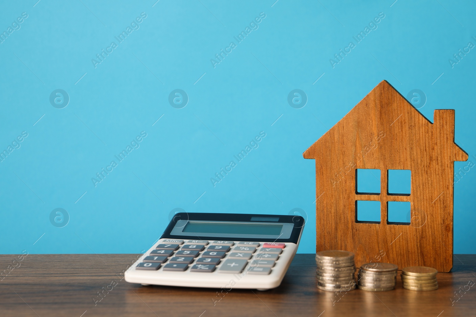 Photo of House model, calculator and stacked coins on wooden table against light blue background, space for text