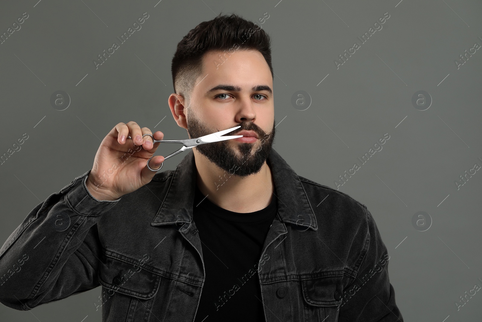 Photo of Handsome young man trimming mustache with scissors on grey background