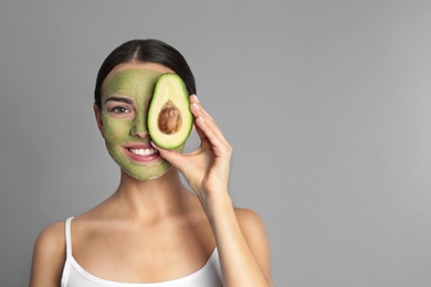Young woman with clay mask on her face holding avocado against grey background, space for text. Skin care