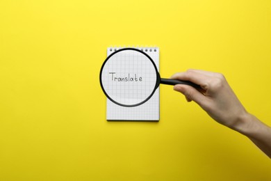 Photo of Woman holding magnifying glass over sheet of paper with word Translate on yellow background, top view