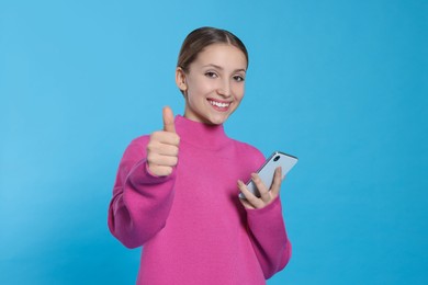 Photo of Teenage girl with smartphone showing thumb up on light blue background