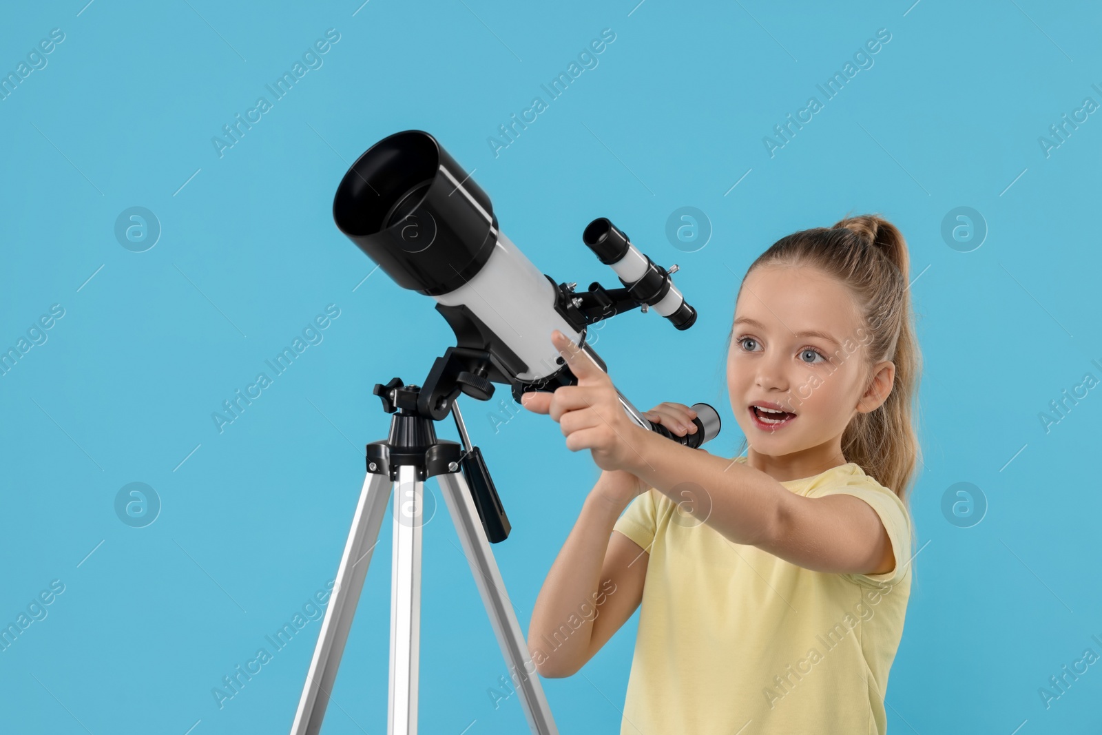 Photo of Excited little girl with telescope pointing at something on light blue background, space for text