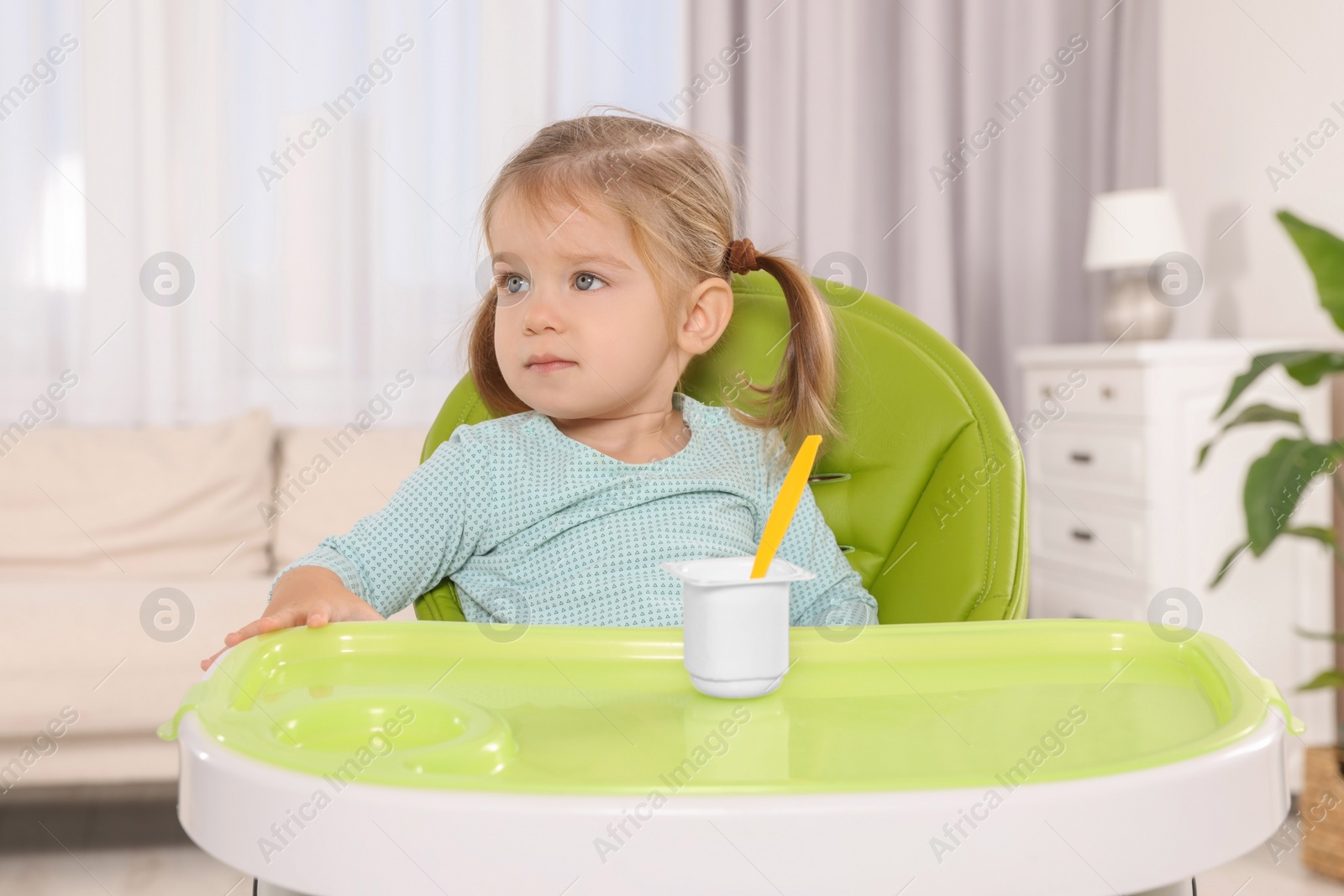 Photo of Cute little child eating tasty yogurt from plastic cup with spoon in high chair indoors