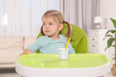 Cute little child eating tasty yogurt from plastic cup with spoon in high chair indoors