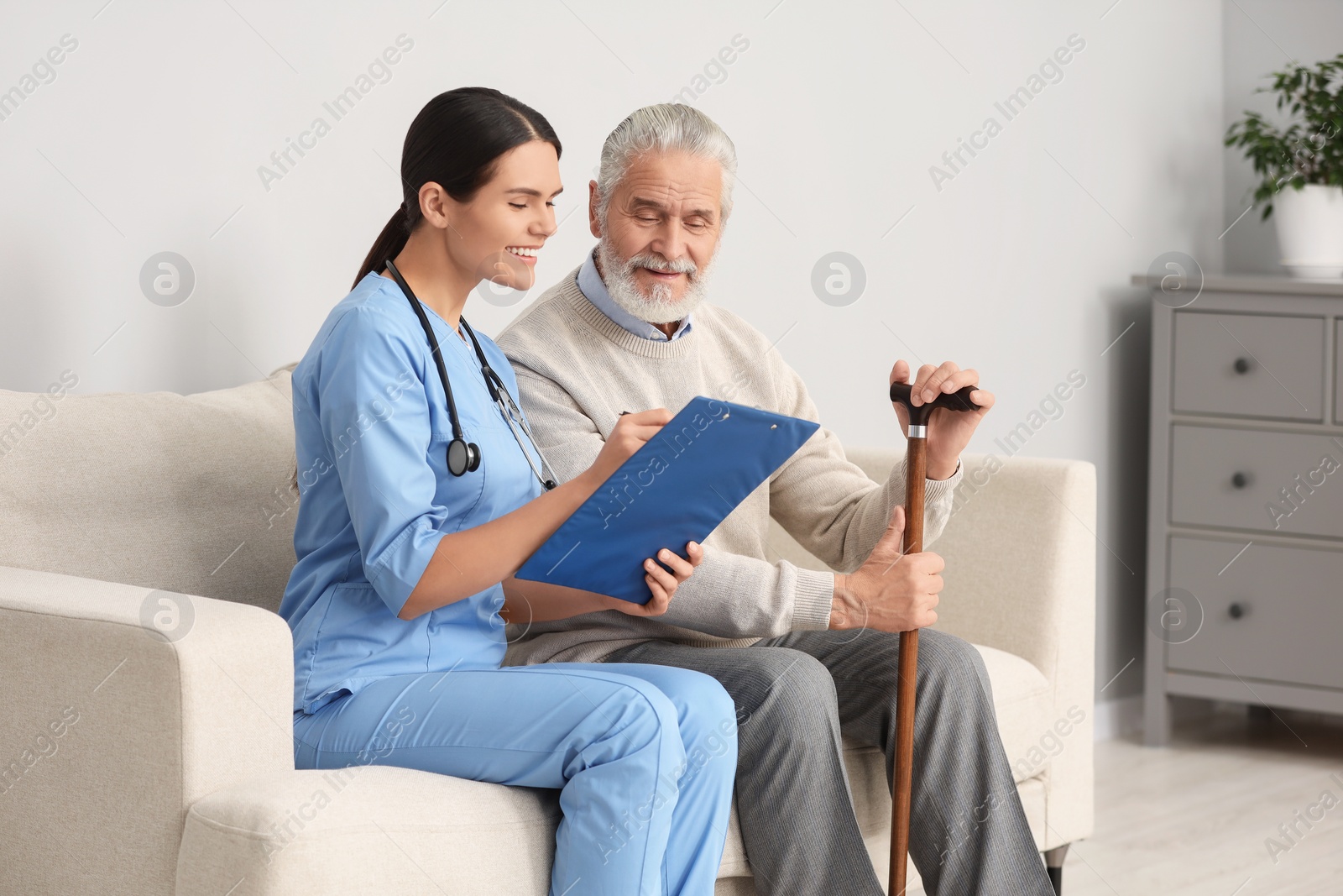 Photo of Smiling nurse with clipboard assisting elderly patient on sofa in hospital