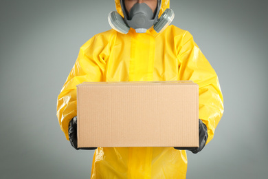 Man wearing chemical protective suit with cardboard box on light grey background, closeup. Prevention of virus spread