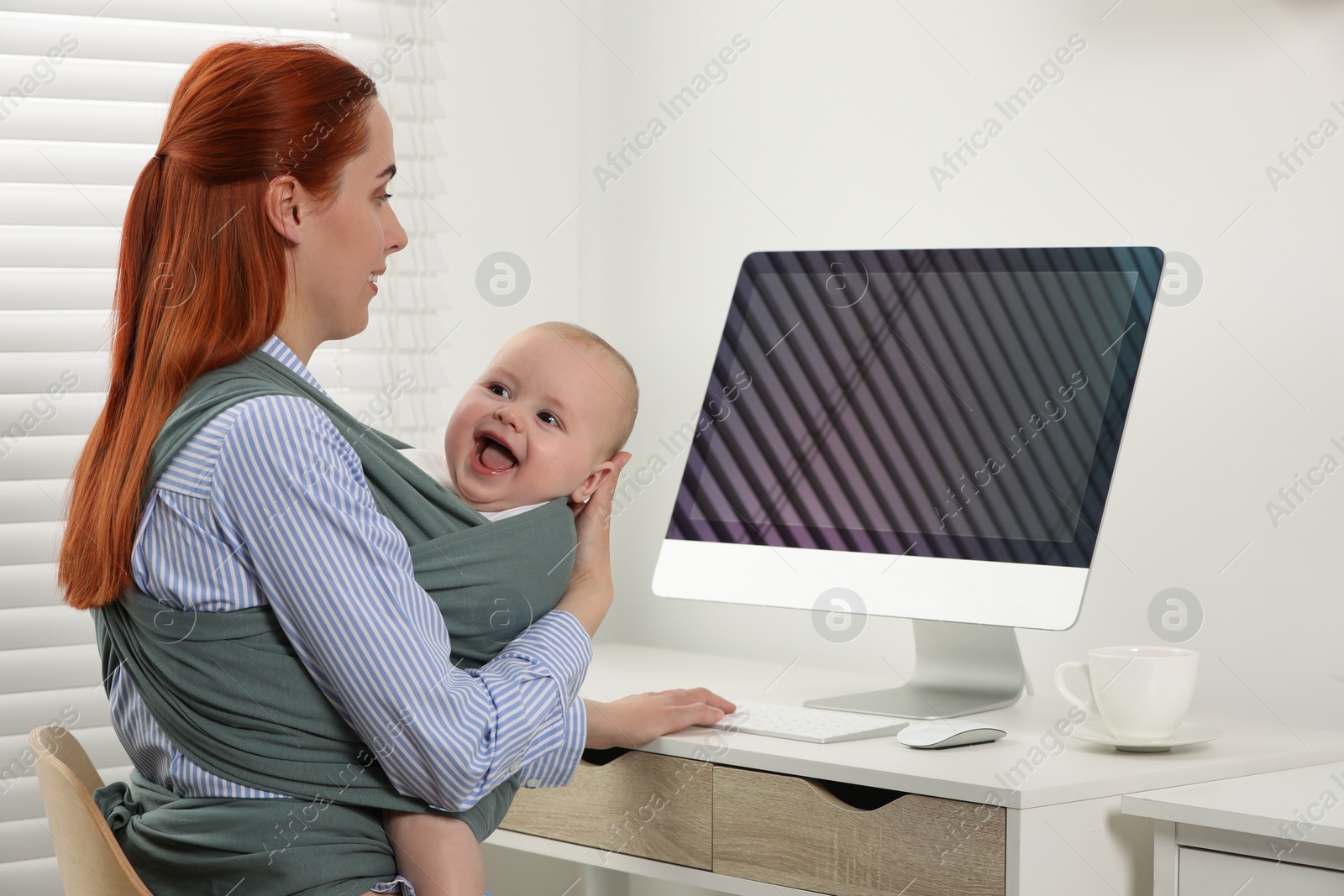 Photo of Mother using computer while holding her child in sling (baby carrier) at workplace