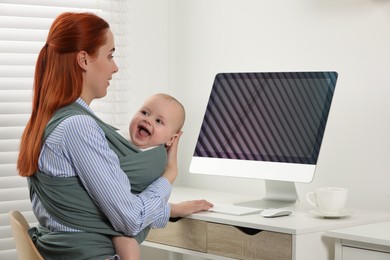 Photo of Mother using computer while holding her child in sling (baby carrier) at workplace