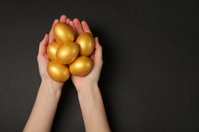 Photo of Woman holding shiny golden eggs on black background, top view. Space for text