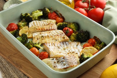 Photo of Pieces of delicious baked cod with vegetables, lemon and spices in dish on wooden table, closeup