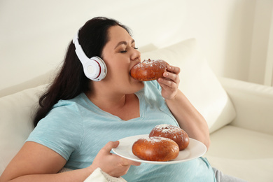Photo of Lazy overweight woman with headphones eating bun at home