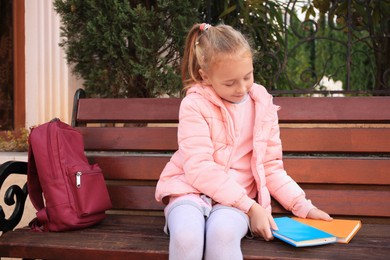 Photo of Cute little girl with backpack and books on bench outdoors
