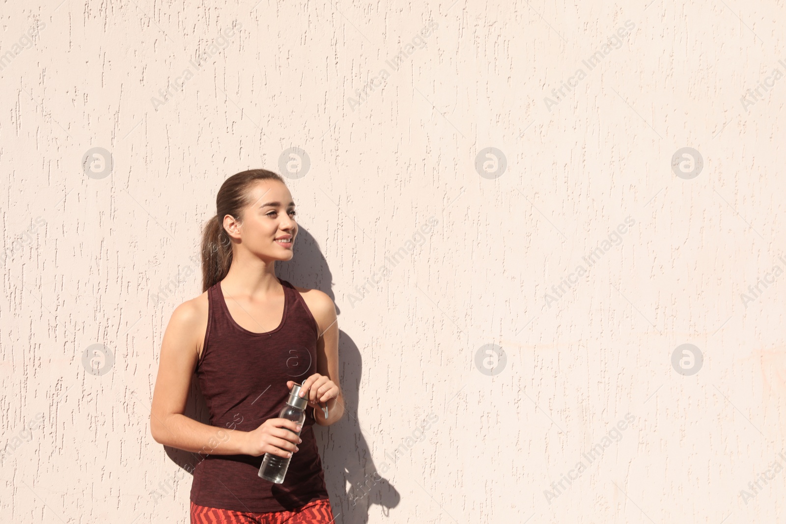 Photo of Young sporty woman holding bottle of water near wall outdoors on sunny day. Space for text