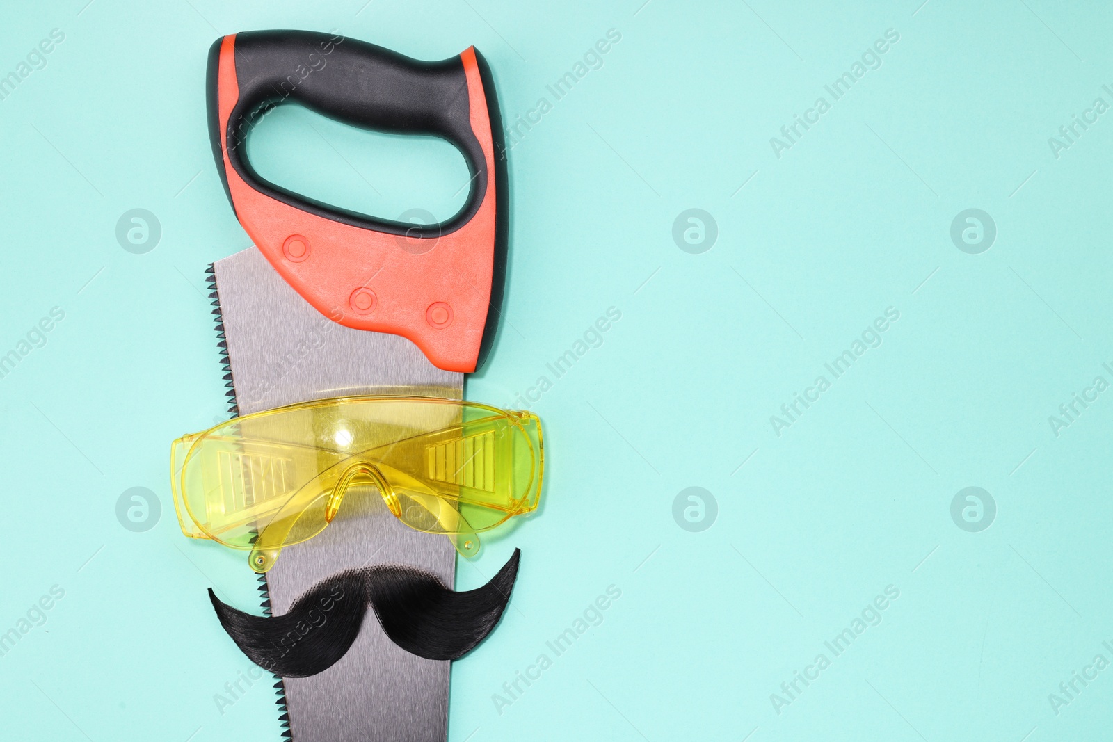 Photo of Man's face made of artificial mustache, safety glasses and hand saw on light blue background, top view. Space for text