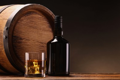 Photo of Whiskey with ice cubes in glass, bottle and barrel on wooden table against black background, space for text