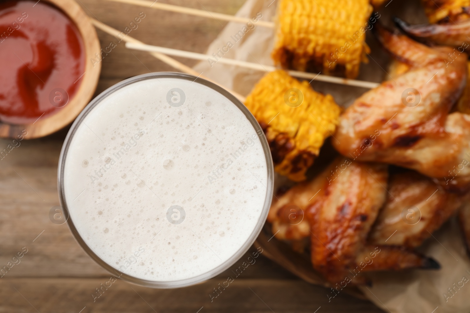 Photo of Glass of beer, delicious baked chicken wings, grilled corn and sauce on wooden table, closeup. Space for text