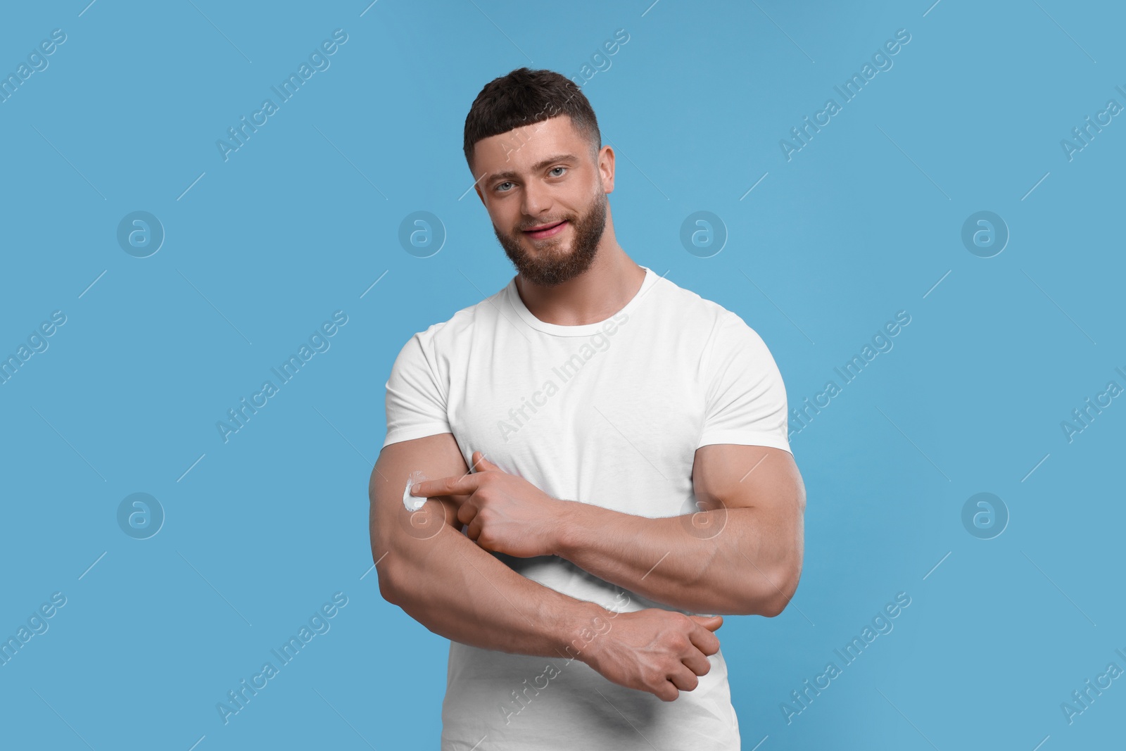 Photo of Handsome man applying body cream onto his arm on light blue background