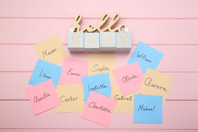 Photo of Cubes with phrase Hello Baby and paper stickers with different names on pink wooden background, flat lay. Choosing baby's name