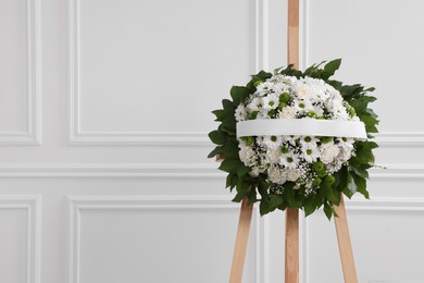 Photo of Funeral wreath of flowers with ribbon on wooden stand near white wall indoors, space for text
