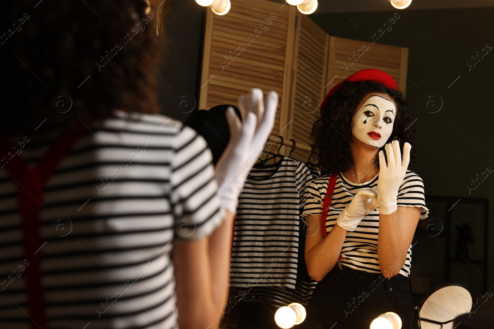 Photo of Young woman in mime costume putting gloves near mirror indoors