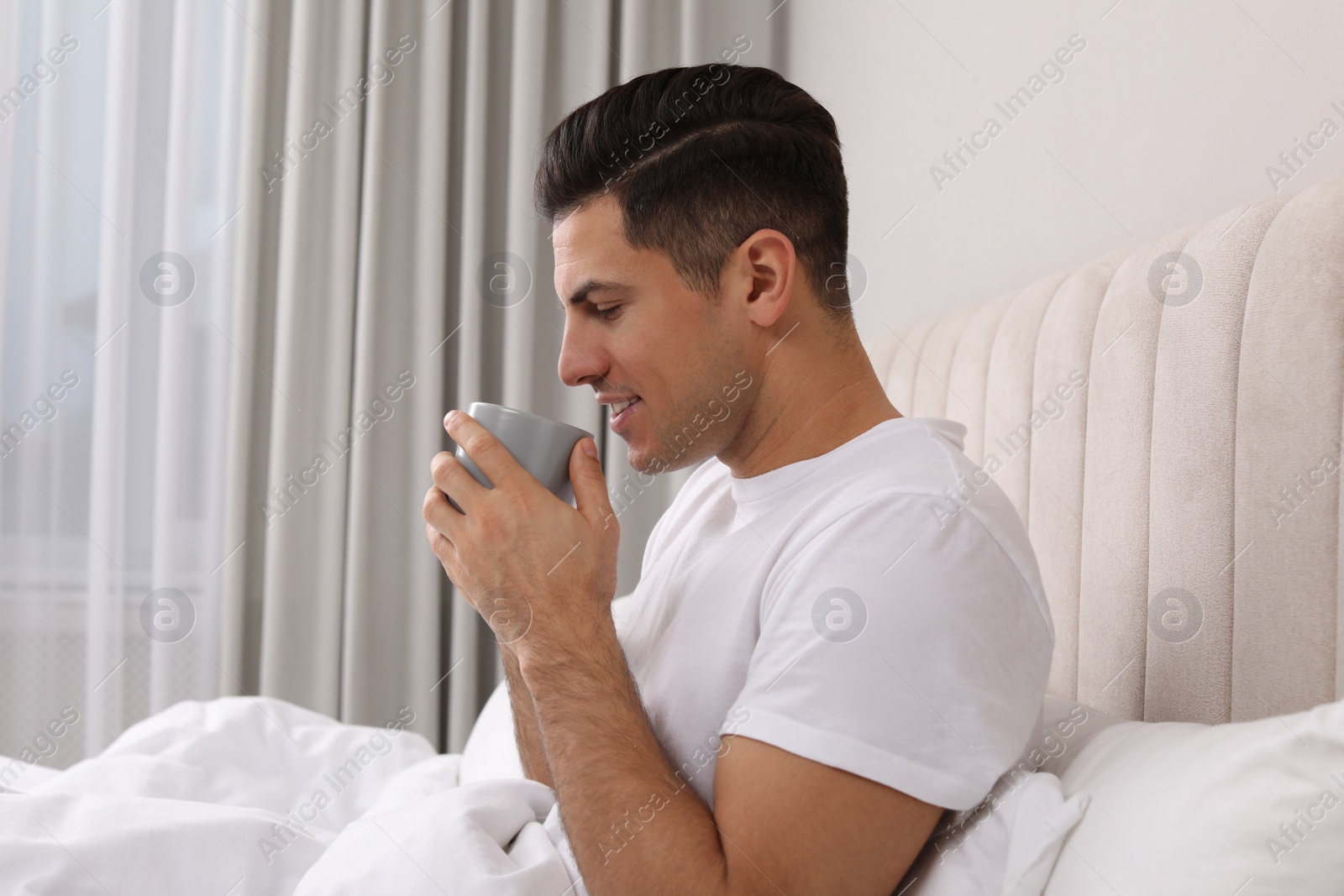 Photo of Happy man with cup of hot drink in bed at home