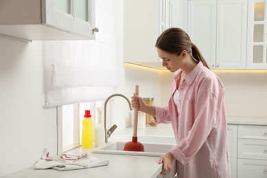 Photo of Young woman using plunger to unclog sink drain in kitchen