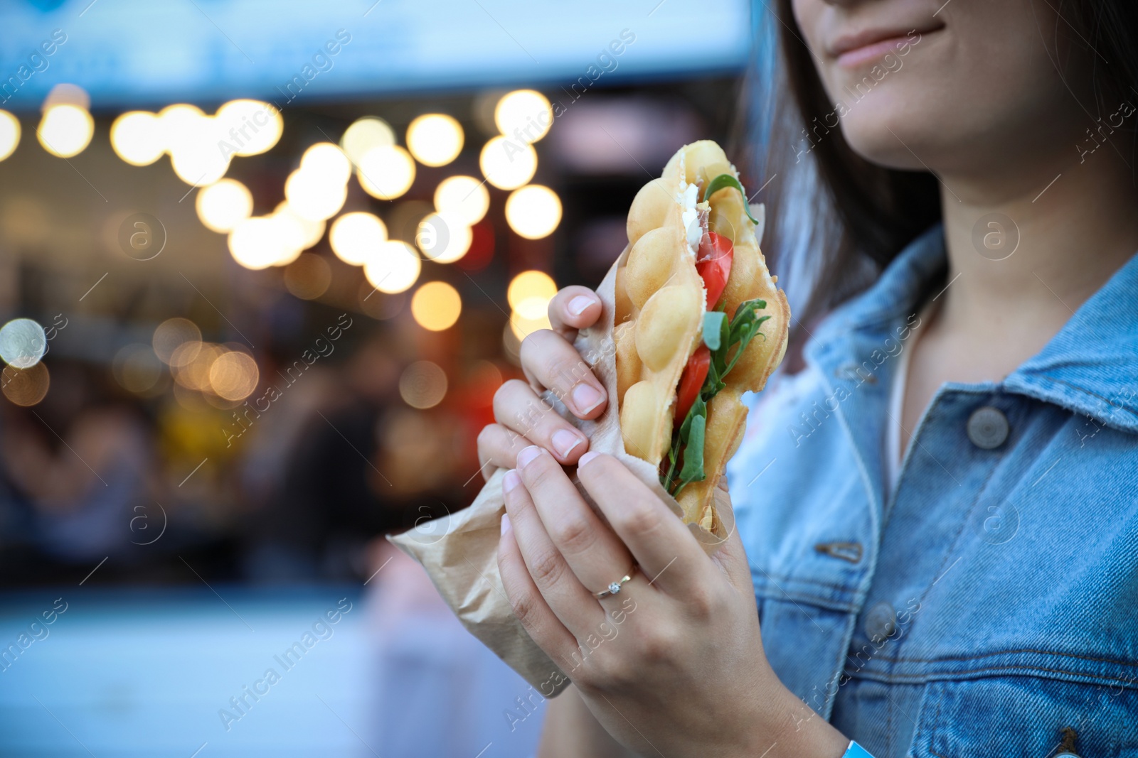 Photo of Young woman holding delicious bubble waffle with tomato and arugula outdoors, closeup