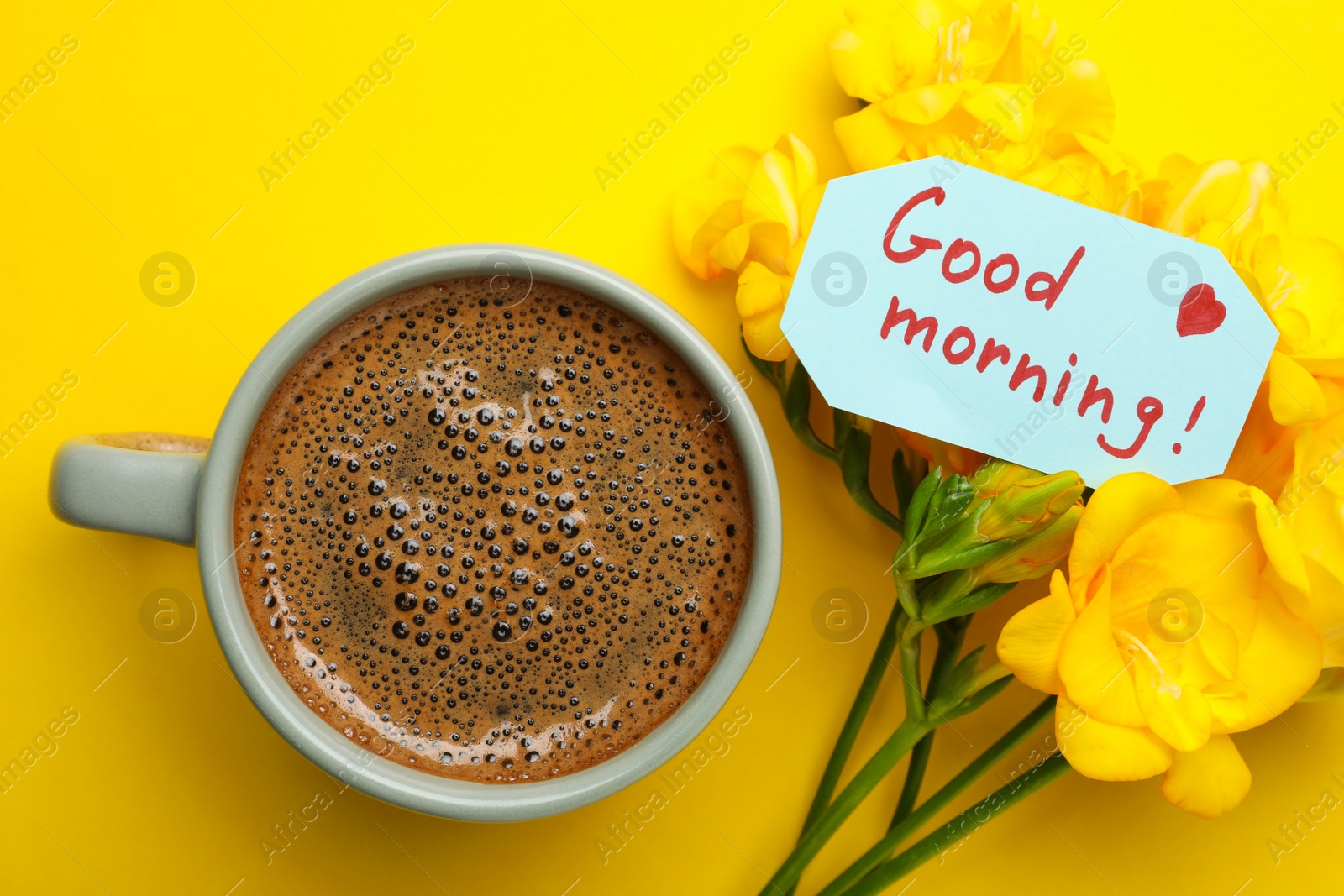 Photo of Cup of aromatic coffee, beautiful freesias and Good Morning note on yellow background, flat lay