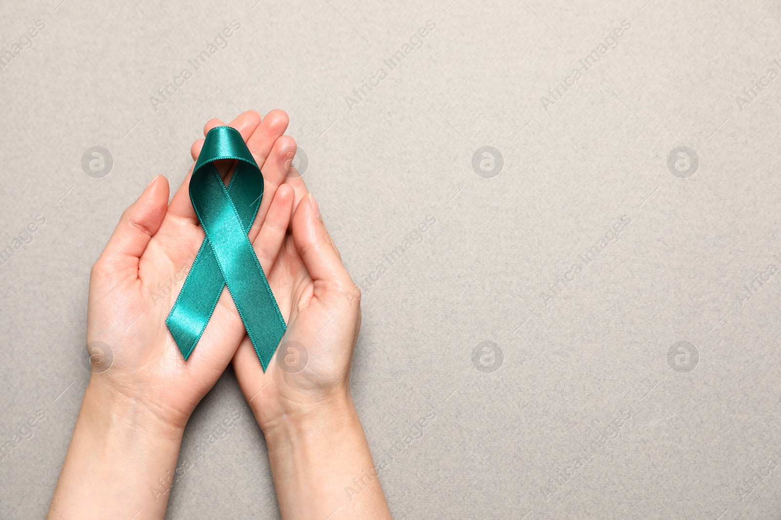 Photo of Woman holding teal awareness ribbon on grey paper, top view with space for text. Symbol of social and medical issues