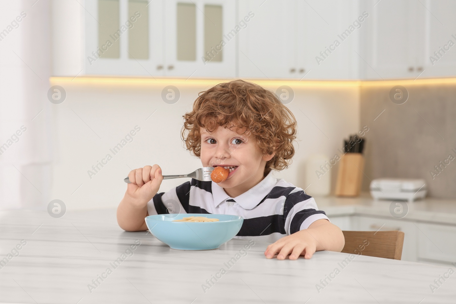 Photo of Cute little boy eating sausage and pasta at table in kitchen