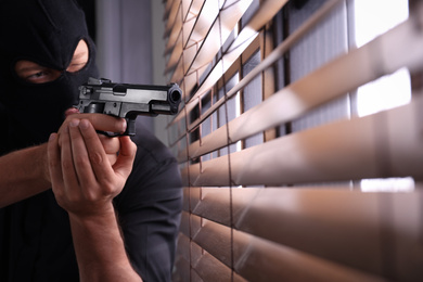 Photo of Man in mask aiming through window blinds indoors, focus on hands