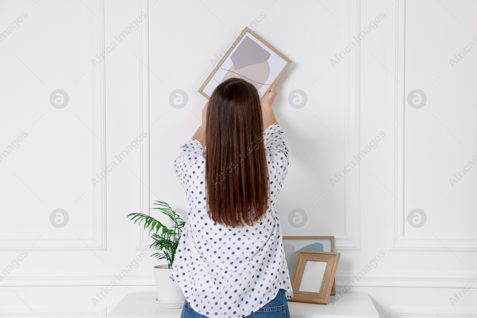 Photo of Woman hanging picture frame on white wall indoors, back view