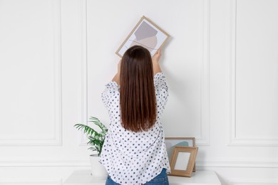 Woman hanging picture frame on white wall indoors, back view