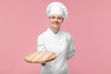 Photo of Happy chef in uniform holding empty wooden board on pink background