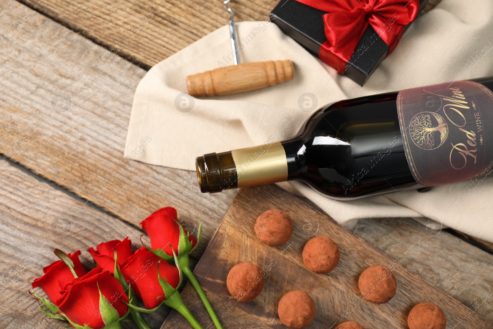 Photo of Bottle of red wine, chocolate truffles, corkscrew, gift box and roses on wooden table, above view