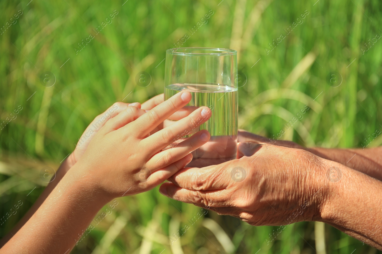 Photo of Child giving glass of water to elderly woman outdoors on sunny day, closeup