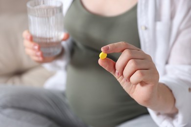 Photo of Pregnant woman holding pill and glass of water on blurred background, closeup