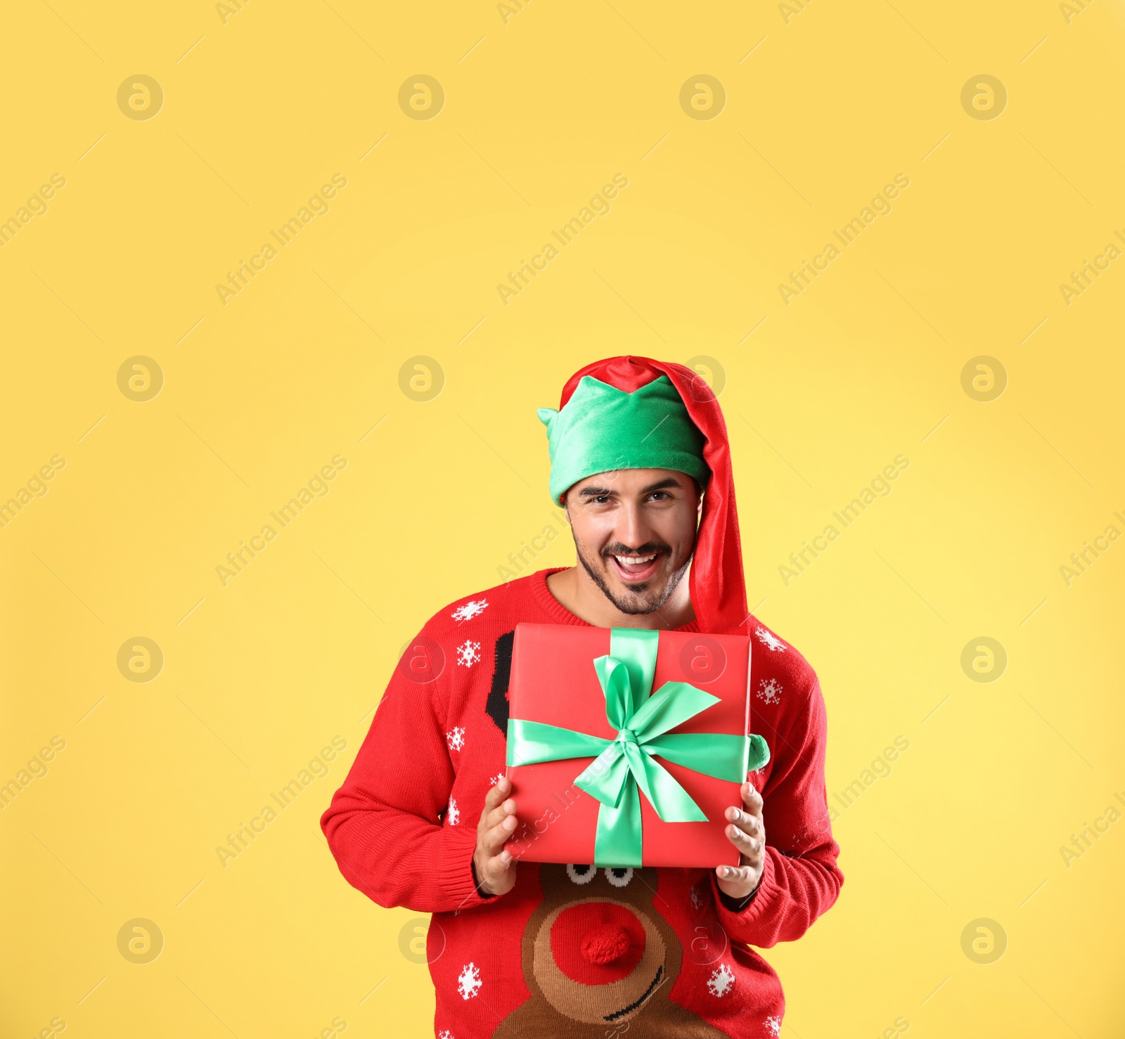 Photo of Happy man in Christmas sweater and elf hat holding gift box on yellow background