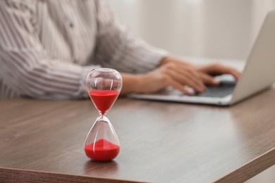 Photo of Hourglass with red flowing sand on table. Woman using laptop indoors, closeup