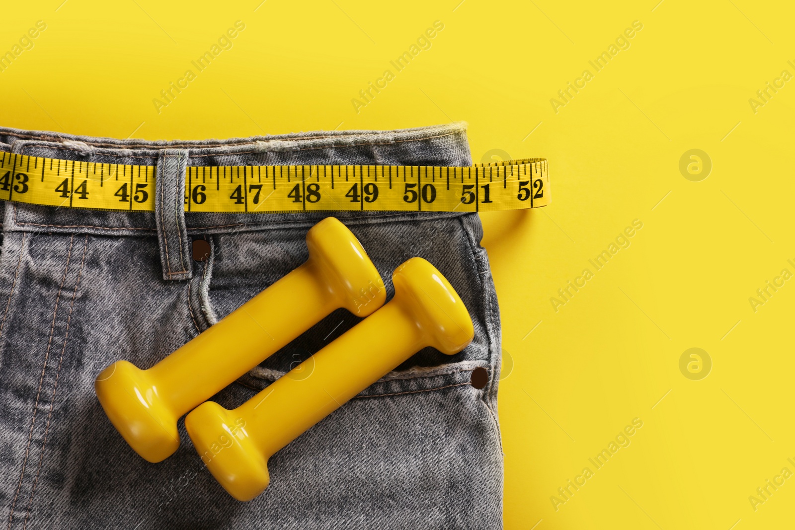 Photo of Jeans with measuring tape, dumbbells on yellow background, top view and space for text. Weight loss concept