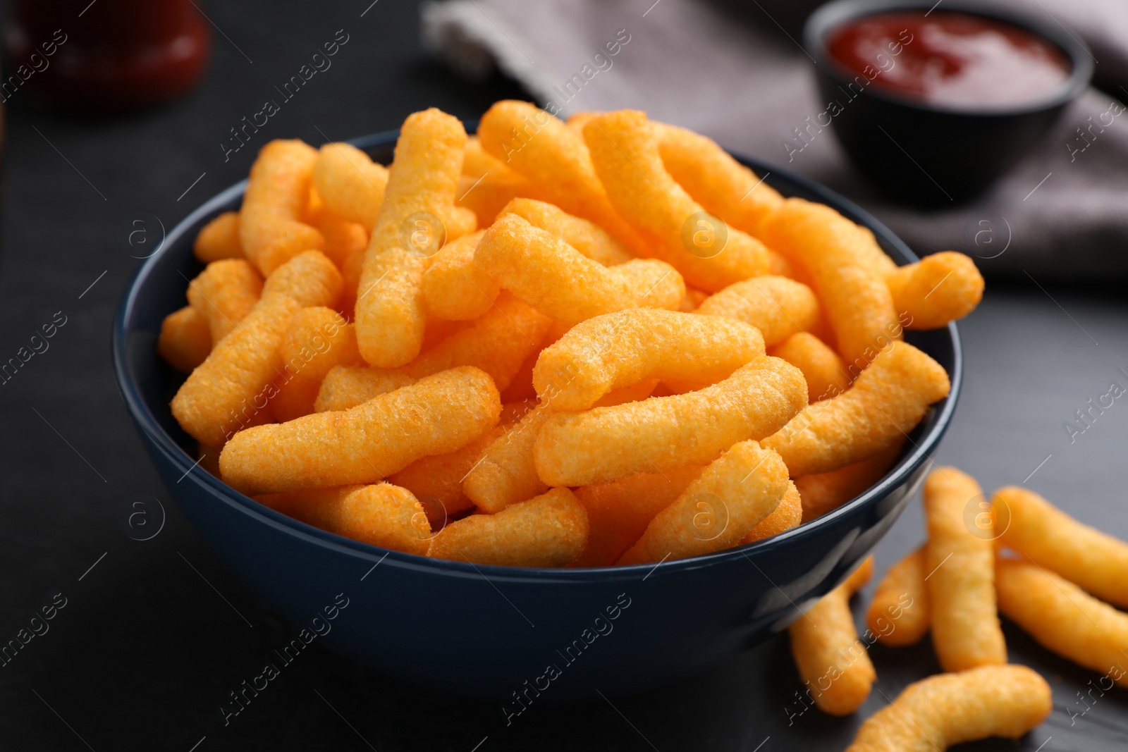 Photo of Bowl with crunchy cheesy corn snack on black table, closeup